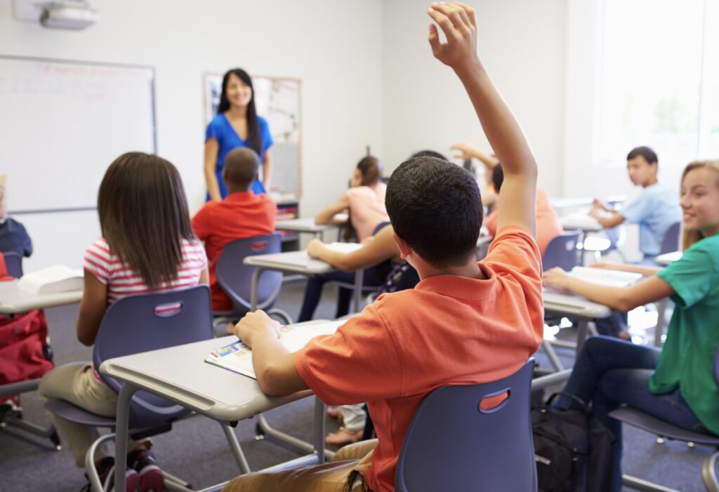 In a classroom, a teacher stands in front next to a white board. A student has his hand raised.